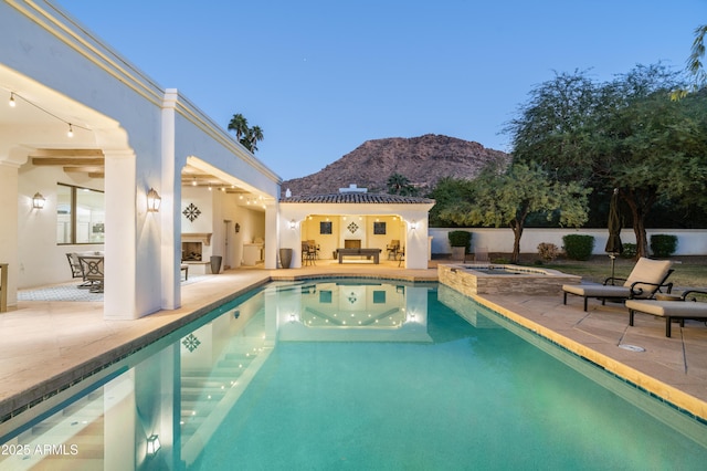 pool at dusk with an outdoor fireplace, a mountain view, an in ground hot tub, and a patio