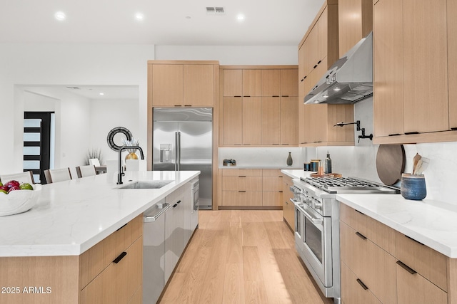 kitchen with modern cabinets, a sink, premium appliances, under cabinet range hood, and light brown cabinets