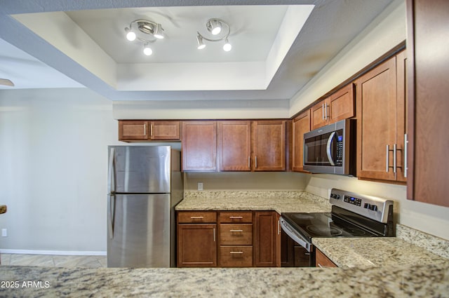 kitchen with stainless steel appliances, baseboards, light stone countertops, a tray ceiling, and brown cabinetry