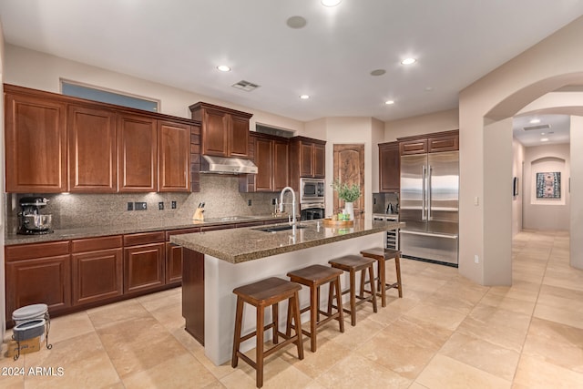 kitchen with sink, a kitchen island with sink, backsplash, built in appliances, and a kitchen breakfast bar