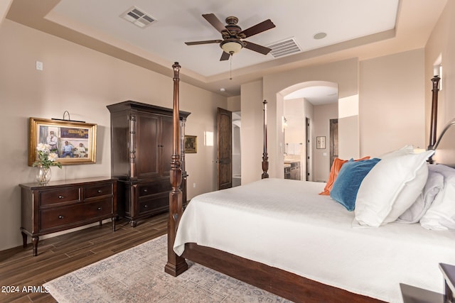 bedroom with ceiling fan, a tray ceiling, ensuite bath, and dark hardwood / wood-style floors