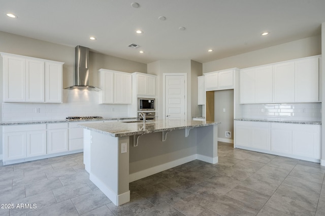 kitchen with sink, white cabinetry, an island with sink, wall chimney range hood, and built in microwave