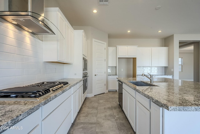 kitchen featuring sink, wall chimney exhaust hood, an island with sink, light stone countertops, and appliances with stainless steel finishes