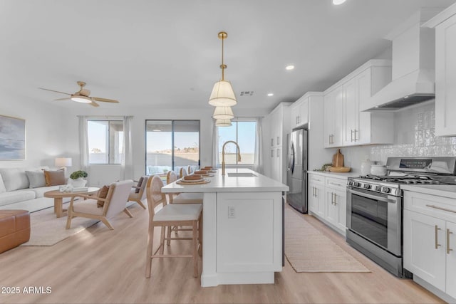 kitchen with wall chimney exhaust hood, white cabinetry, decorative light fixtures, an island with sink, and stainless steel appliances