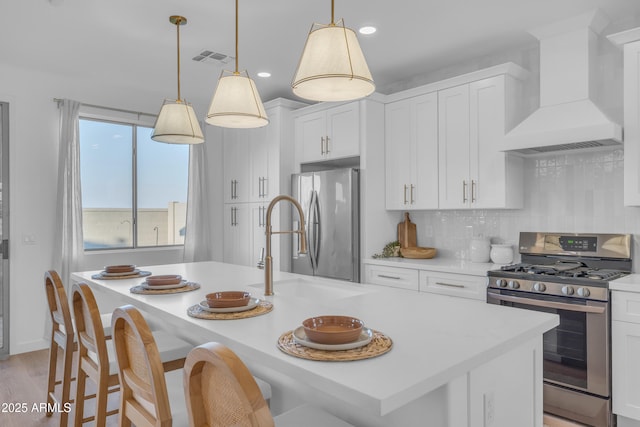 kitchen with white cabinetry, appliances with stainless steel finishes, custom range hood, pendant lighting, and a kitchen island with sink