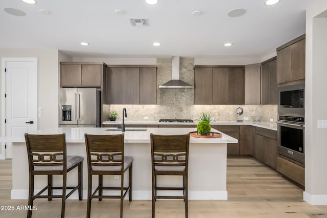 kitchen with a center island with sink, wall chimney range hood, light countertops, a sink, and stainless steel appliances