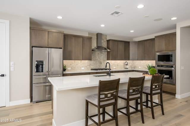 kitchen with wall chimney range hood, dark brown cabinets, a sink, stainless steel appliances, and visible vents
