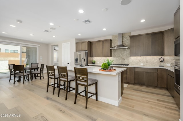 kitchen with decorative backsplash, a center island with sink, light countertops, wall chimney exhaust hood, and appliances with stainless steel finishes