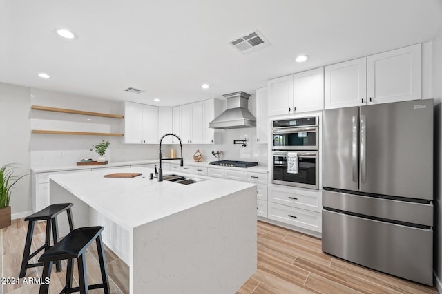 kitchen with sink, wall chimney range hood, a kitchen island with sink, white cabinets, and appliances with stainless steel finishes