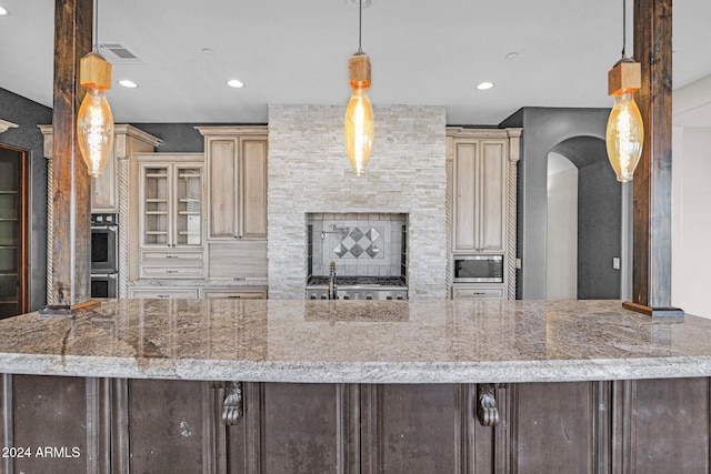 kitchen with light stone countertops, stainless steel appliances, a stone fireplace, cream cabinets, and pendant lighting