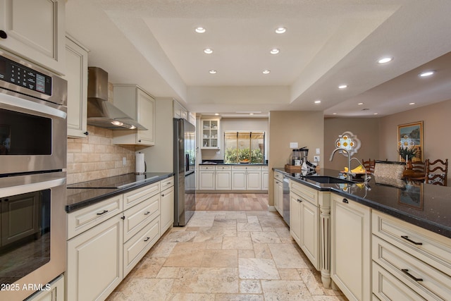 kitchen with recessed lighting, appliances with stainless steel finishes, cream cabinetry, wall chimney exhaust hood, and stone tile flooring
