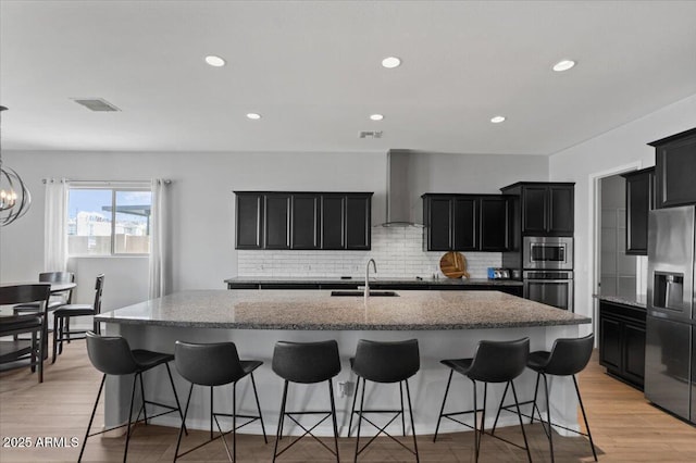 kitchen with a spacious island, visible vents, stainless steel appliances, wall chimney exhaust hood, and dark cabinets