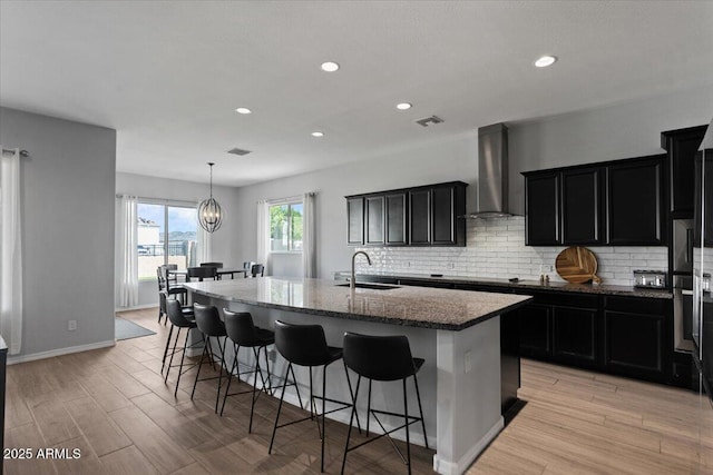 kitchen featuring a breakfast bar, an island with sink, a sink, wall chimney range hood, and tasteful backsplash