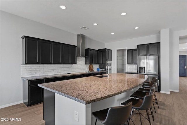 kitchen featuring a sink, a spacious island, dark cabinetry, stainless steel appliances, and wall chimney range hood