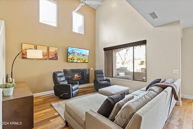 living room with ceiling fan, light wood-type flooring, a fireplace, and a high ceiling