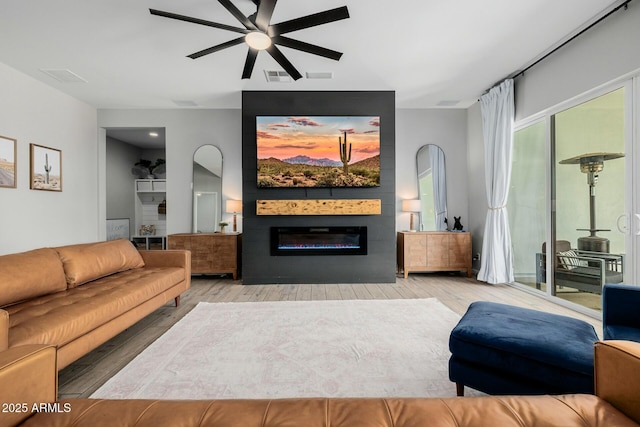 living area with a ceiling fan, visible vents, wood finished floors, and a glass covered fireplace