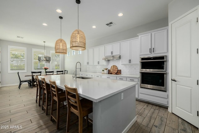 kitchen with decorative backsplash, double oven, a sink, light wood-type flooring, and under cabinet range hood