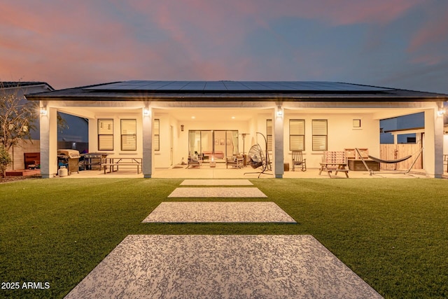 back of property at dusk with a yard, roof mounted solar panels, a patio, and stucco siding