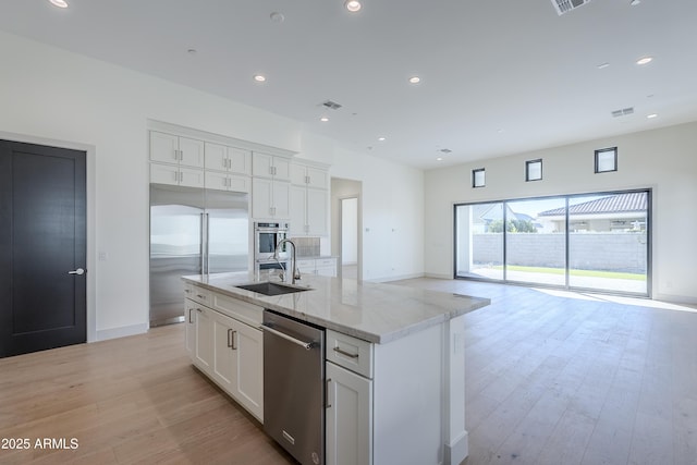 kitchen featuring appliances with stainless steel finishes, a sink, light stone countertops, a kitchen island with sink, and white cabinets