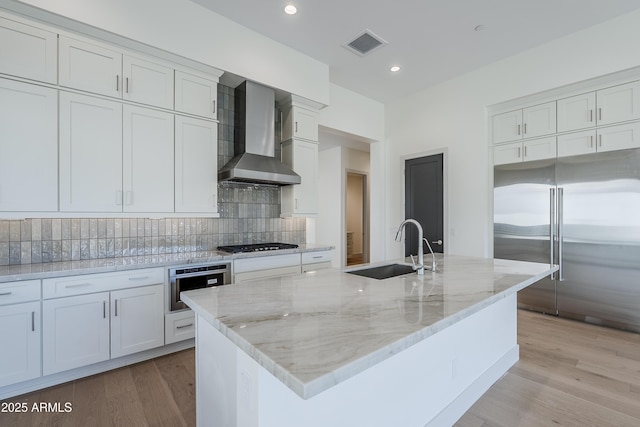 kitchen featuring wall chimney exhaust hood, stainless steel appliances, a sink, decorative backsplash, and light wood-style flooring