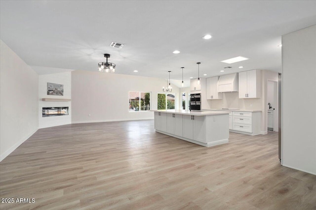 kitchen with white cabinetry, double oven, custom range hood, pendant lighting, and a kitchen island with sink