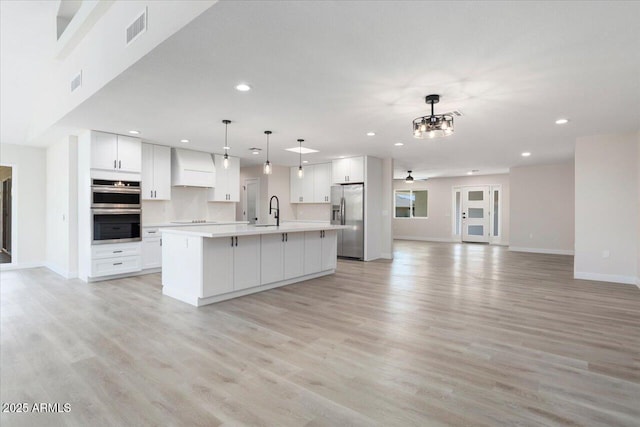 kitchen featuring decorative light fixtures, an island with sink, white cabinetry, stainless steel appliances, and wall chimney range hood