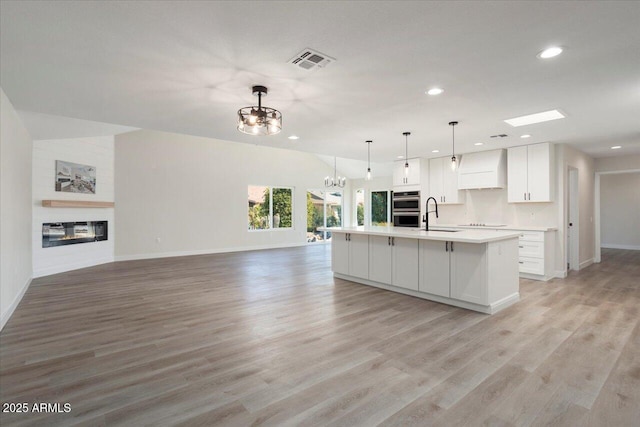 kitchen featuring decorative light fixtures, double oven, custom range hood, a kitchen island with sink, and white cabinets