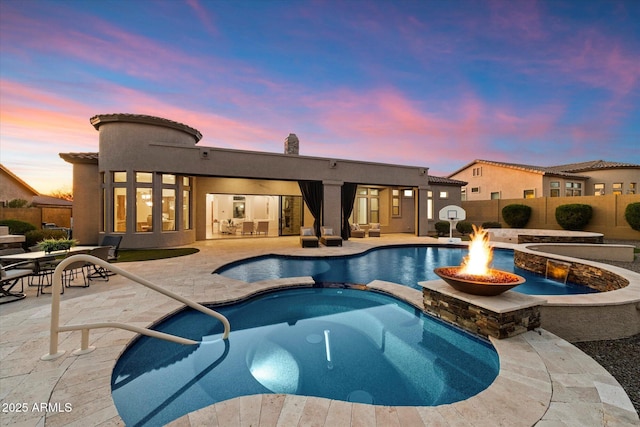 pool at dusk featuring a patio, a fenced backyard, a pool with connected hot tub, and a fire pit