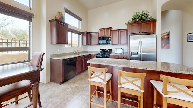 kitchen featuring black appliances, a sink, a breakfast bar area, light stone countertops, and a towering ceiling