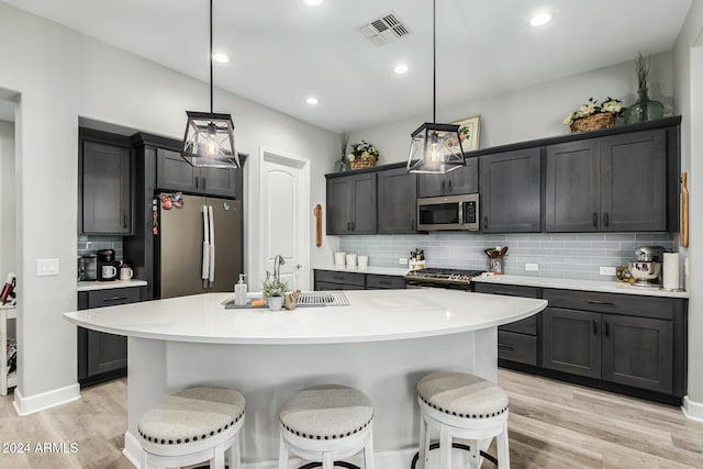 kitchen featuring light wood-type flooring, appliances with stainless steel finishes, decorative light fixtures, and a kitchen island with sink