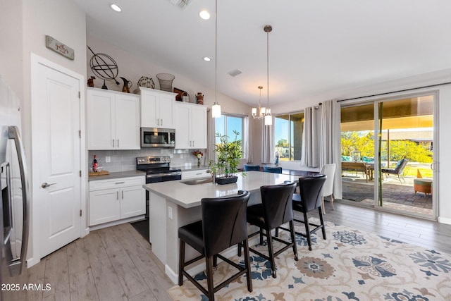 kitchen with tasteful backsplash, stainless steel appliances, white cabinets, hanging light fixtures, and an island with sink