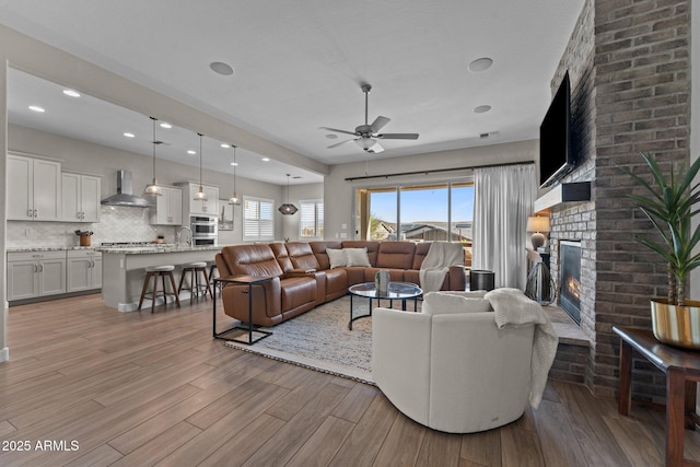 living room with ceiling fan, a brick fireplace, light wood-style flooring, and recessed lighting