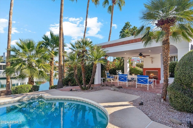 view of pool with ceiling fan, a water view, a patio, and exterior fireplace