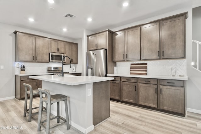 kitchen with an island with sink, appliances with stainless steel finishes, light hardwood / wood-style flooring, and decorative backsplash