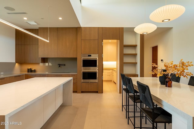 kitchen featuring light stone countertops, a large island, brown cabinetry, stainless steel double oven, and modern cabinets