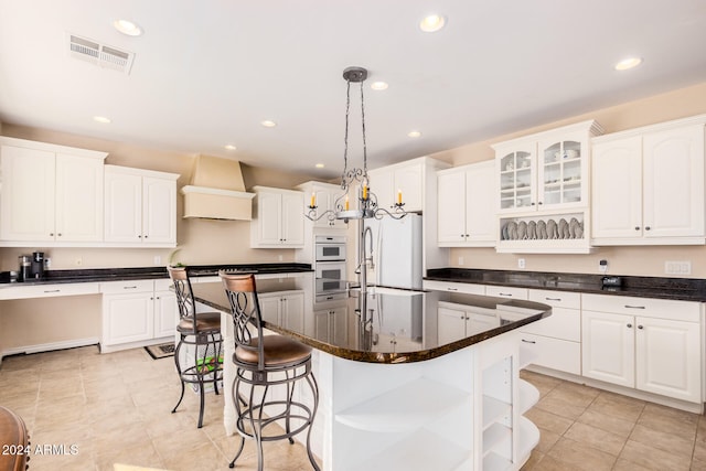 kitchen with white appliances, hanging light fixtures, white cabinetry, custom exhaust hood, and a kitchen island with sink