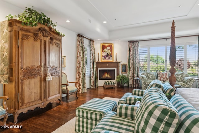 living area featuring a warm lit fireplace, a raised ceiling, dark wood-style flooring, and recessed lighting