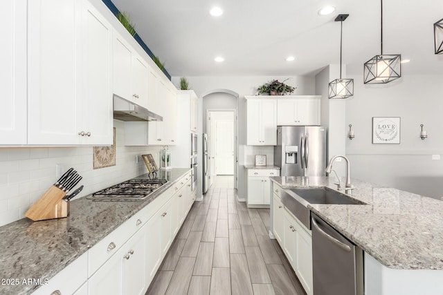 kitchen featuring a kitchen island with sink, under cabinet range hood, stainless steel appliances, arched walkways, and decorative backsplash