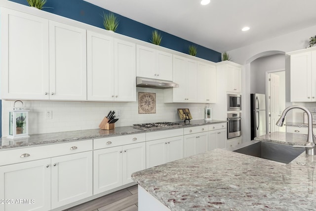 kitchen with a sink, under cabinet range hood, arched walkways, appliances with stainless steel finishes, and white cabinets