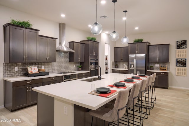 kitchen with decorative light fixtures, sink, wall chimney range hood, stainless steel appliances, and a center island with sink