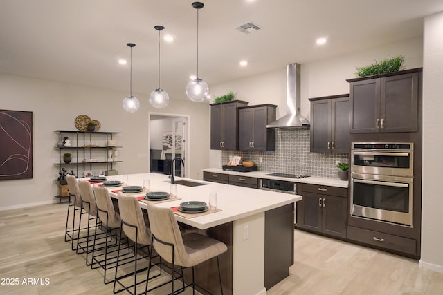 kitchen with wall chimney exhaust hood, hanging light fixtures, a kitchen breakfast bar, stainless steel double oven, and a kitchen island with sink