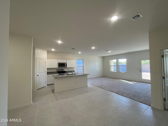 kitchen with appliances with stainless steel finishes, dark stone counters, light colored carpet, white cabinets, and an island with sink