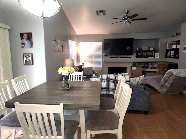 dining room featuring wood finished floors, a ceiling fan, vaulted ceiling, and a tiled fireplace