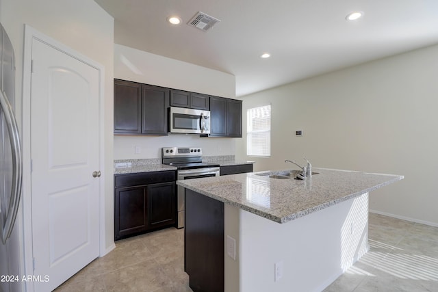 kitchen with stainless steel appliances, a kitchen island with sink, light stone counters, dark brown cabinetry, and sink