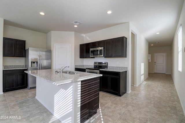 kitchen with appliances with stainless steel finishes, a kitchen island with sink, light stone counters, dark brown cabinetry, and sink