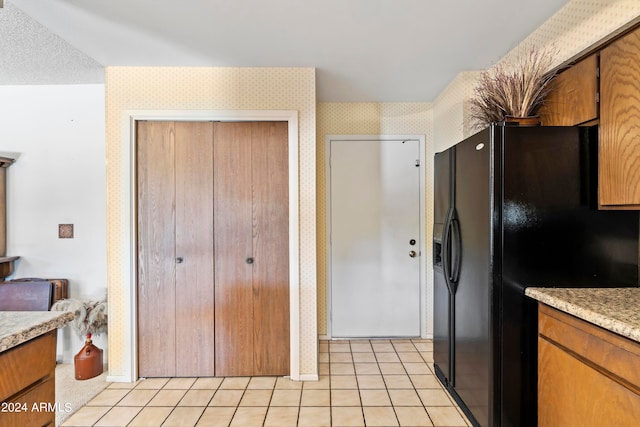 kitchen with wallpapered walls, light tile patterned floors, baseboards, brown cabinetry, and black fridge with ice dispenser