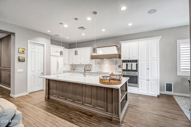 kitchen featuring white cabinets, a center island with sink, paneled refrigerator, decorative light fixtures, and stainless steel double oven