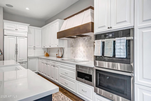 kitchen with premium range hood, black electric stovetop, paneled built in fridge, white cabinetry, and stainless steel double oven