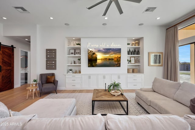 living room with built in shelves, a barn door, ceiling fan, and light wood-type flooring