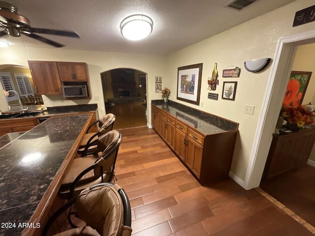 kitchen with arched walkways, brown cabinetry, stainless steel microwave, a kitchen breakfast bar, and light wood-type flooring